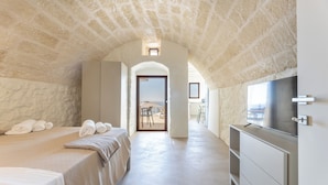 A bedroom with a stone ceiling, a bed with white linens, a TV, and a view through an open door to a balcony.