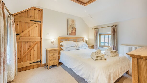 A bedroom with a wooden door, a bed with white linens, a nightstand, and a window with curtains.