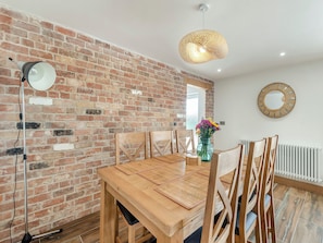 A dining area with a brick wall, wooden table, chairs, a hanging light fixture, and a vase with flowers.