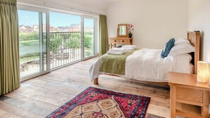 Une chambre à coucher comprenant un lit, une table de chevet en bois, une commode, un miroir et une vue sur l’extérieur, accessible par une porte-fenêtre coulissante.