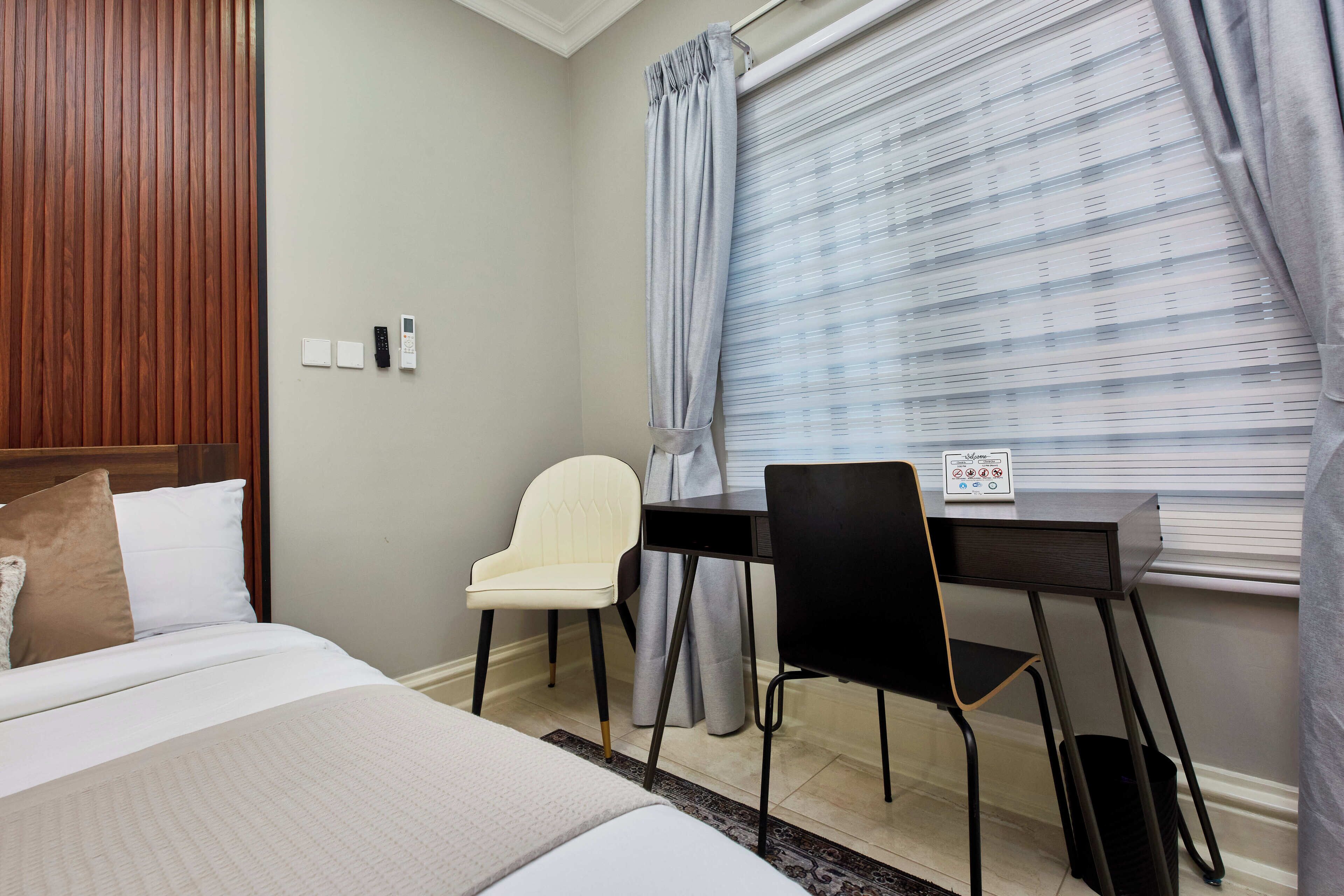 Quarto de hotel com cama, escrivaninha, duas cadeiras, janela com persianas e painel de controle fixado na parede.