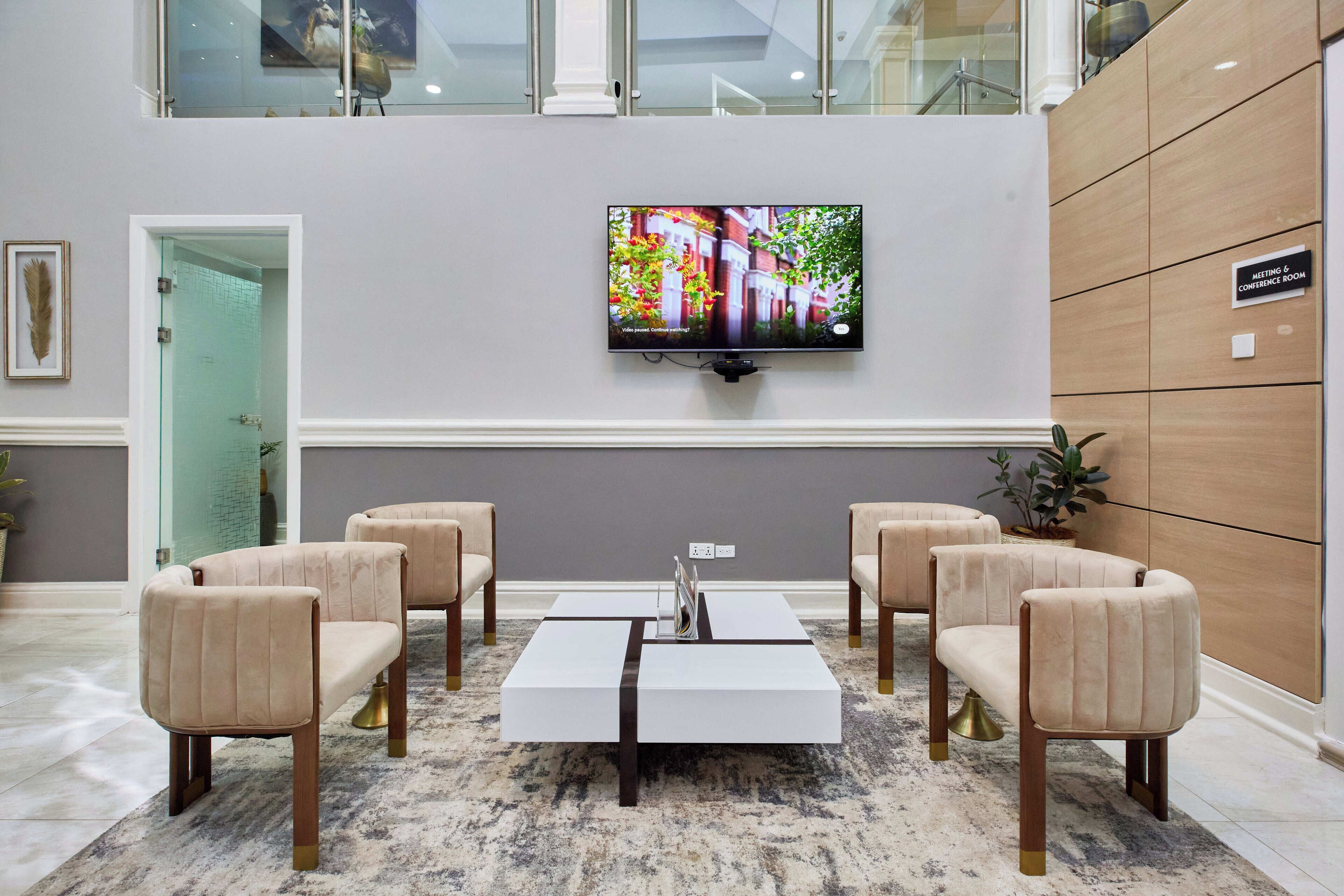 A lobby with a modern reception desk, a flat-screen TV mounted on the wall, and a seating area with four cushioned chairs.