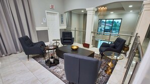 A modern hotel lobby with a seating area featuring two armchairs, a coffee table, and a decorative bowl. A glass railing overlooks a balcony with a view of the outdoors. - The Mayflower Inn (Kwabenya)