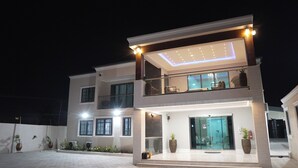 A modern two-story house with a balcony, large windows, and a well-lit entrance. - The Mayflower Inn (Kwabenya)