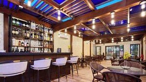 A bar area with wooden beams, a bar counter, and shelves stocked with bottles. There are bar stools and a dining area with chairs and a table. - The Mayflower Inn (Kwabenya)
