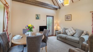 A cozy living room with a dining area, wooden beams on the ceiling, a TV, and a window with curtains. - The Granary - UK44490 (Sedgefield)