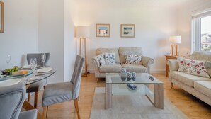 Ein Wohnzimmer mit einem Glastisch, grau gepolsterten Sesseln, einer Couch mit gemusterten Kissen und zwei Stehlampen.