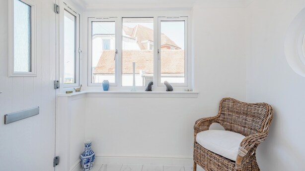 Ein Zimmer mit weißen Wänden, ein Korbstuhl mit Kissen, ein Fenster mit Blick auf Gebäude und eine blaue Vase auf dem Fensterbrett.