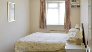 A bedroom with a bed, a bedside table with a lamp, a window with curtains, and electrical outlets on the wall. - Smugglers Haunt - UK44513 (Hope Cove, near Kingsbridge)