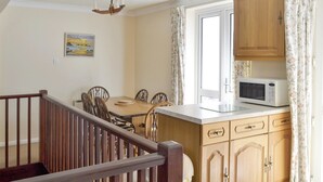 A kitchen with wooden cabinets, a dining table with chairs, a microwave, and a window with curtains. - Smugglers Haunt - UK44513 (Hope Cove, near Kingsbridge)
