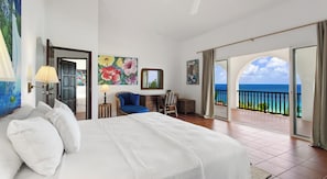 Habitación de hotel con cama, una silla, un escritorio, un cuadro y balcón con vista al océano.