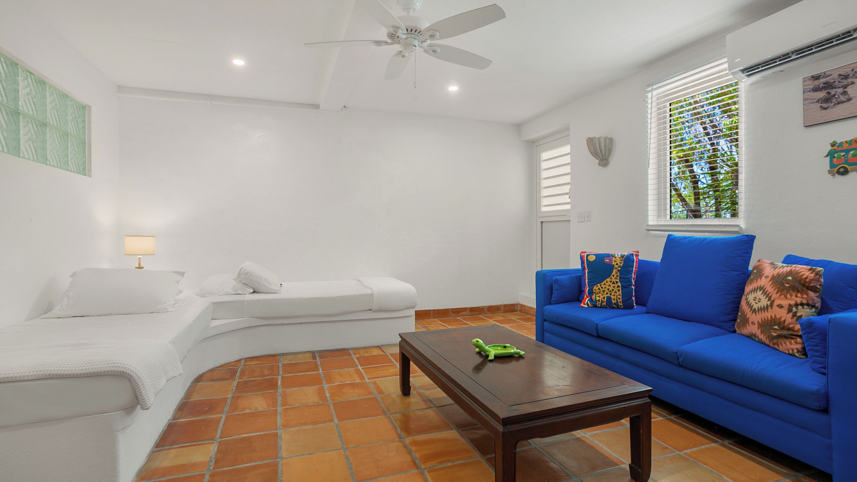 Una sala de estar con un sofá blanco, un sofá azul con cojines estampados, una mesa de centro de madera, un ventilador de techo y una ventana con persianas.