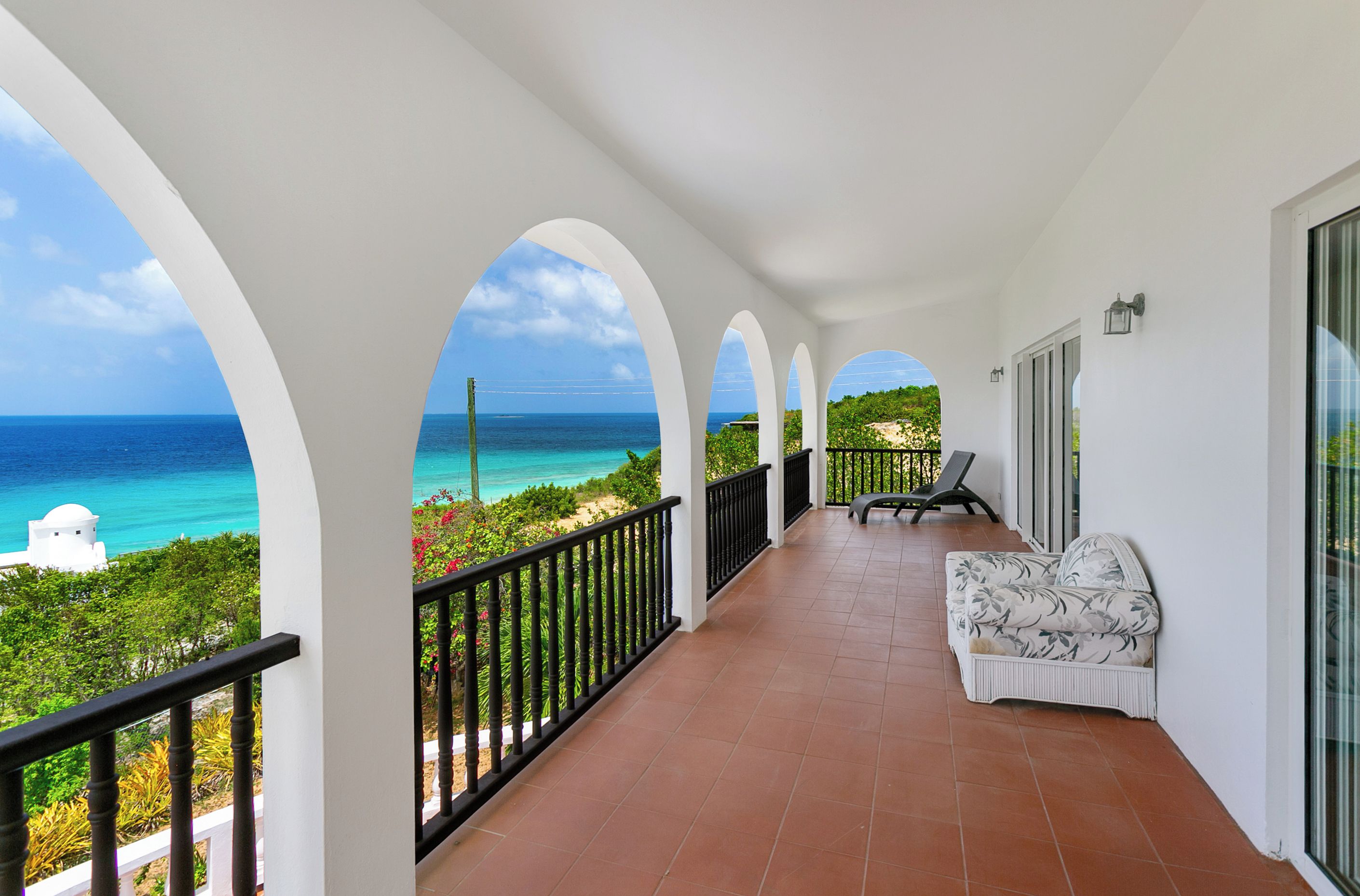 A covered balcony with a view of the ocean, a white building, and a chair.