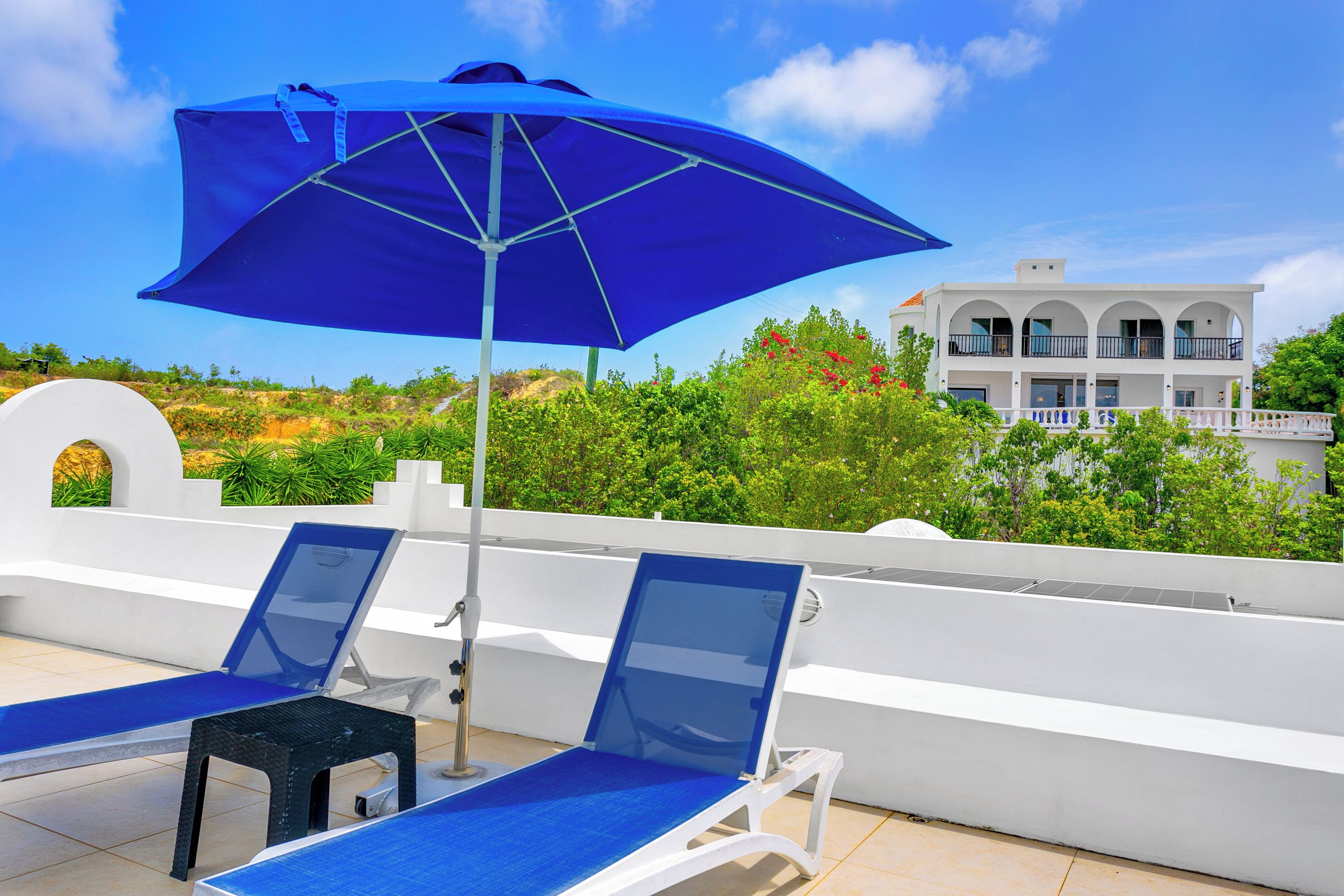 A rooftop terrace with two lounge chairs and a blue umbrella, overlooking a white building and lush greenery.