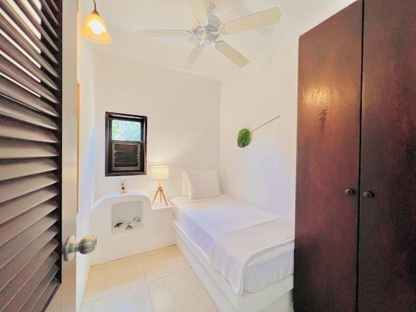Una habitación pequeña y limpia con cama, mesita de noche con lámpara, ventana y ventilador de techo.