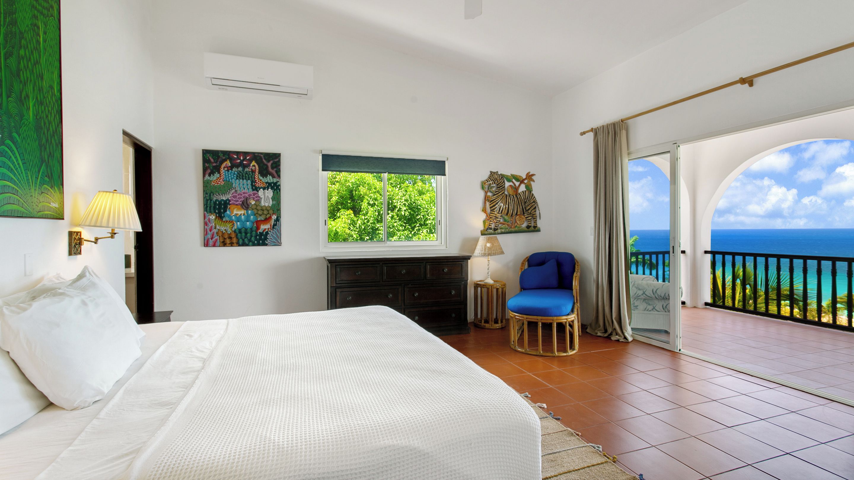 A bedroom with a bed, a dresser, a chair, a window with a view, and a balcony with a railing.