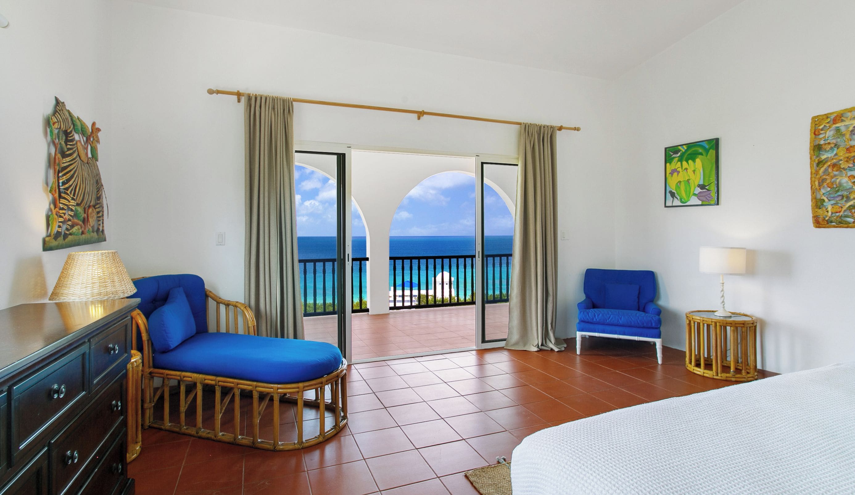 A hotel room with a bed, two blue chairs, a wooden dresser, a lamp, and a view of the ocean.