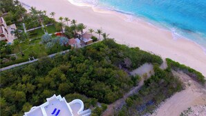 Vista aérea de un complejo turístico en la playa, con una costa arenosa, palmeras y algunos edificios que incluyen una piscina.