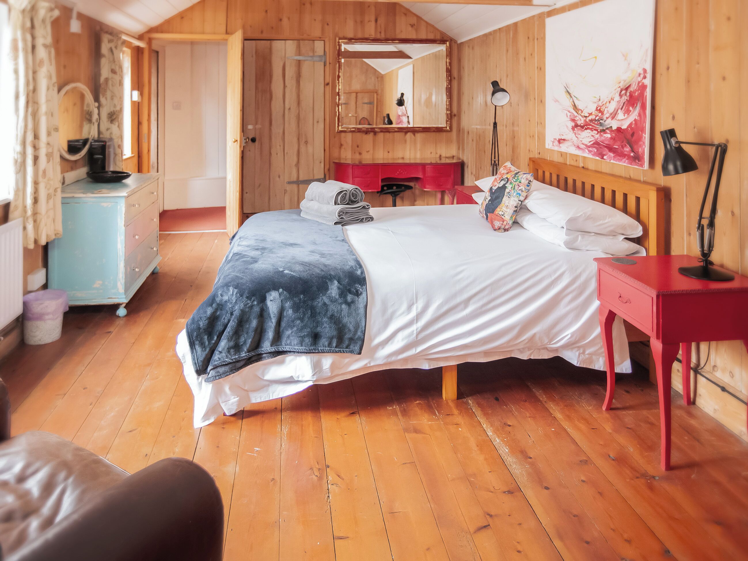 A wooden-paneled room with a bed, a red nightstand, a mirror, and a painting on the wall.