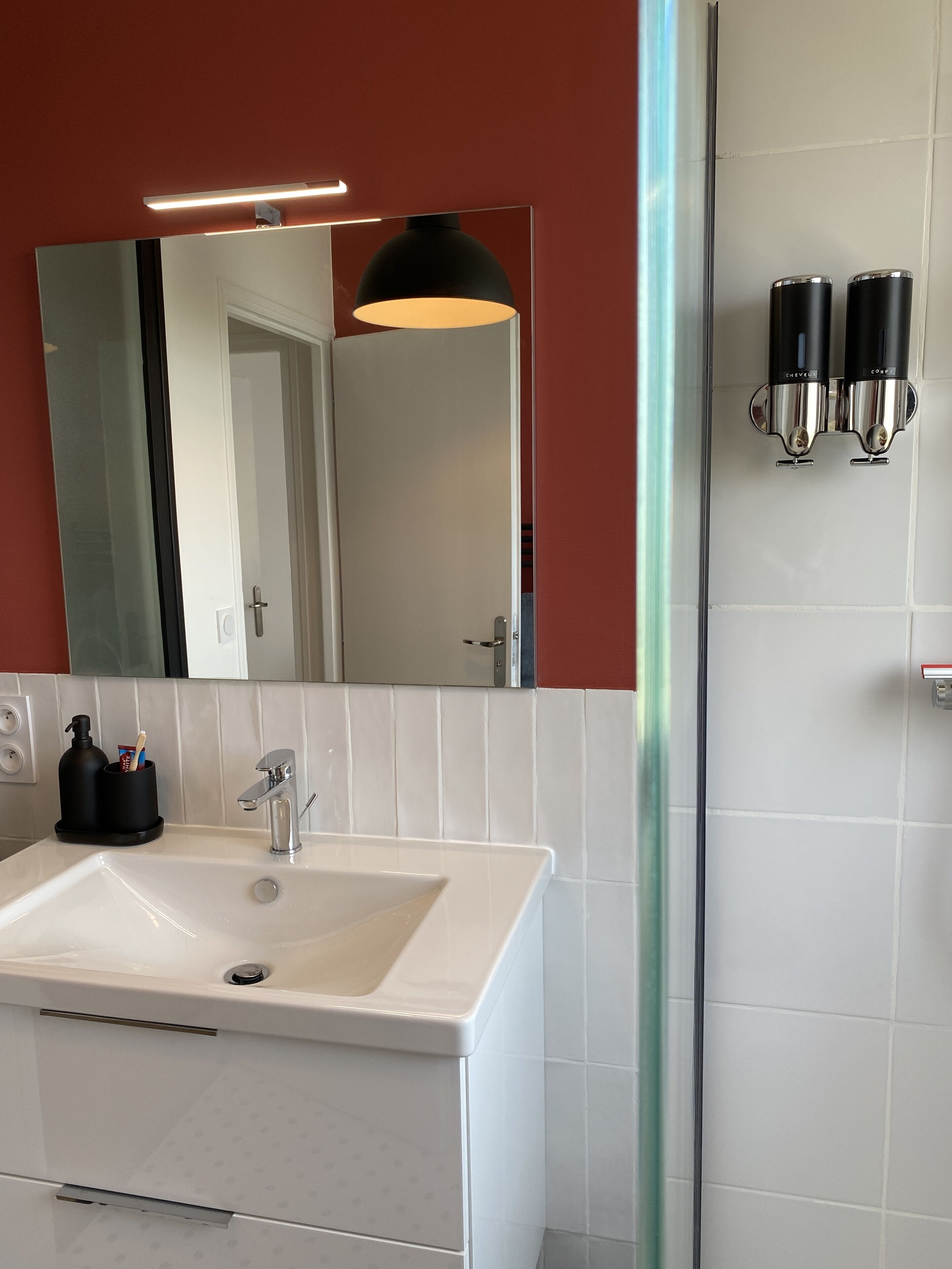 A bathroom with a white sink, a mirror, a black pendant light, and two black soap dispensers.
