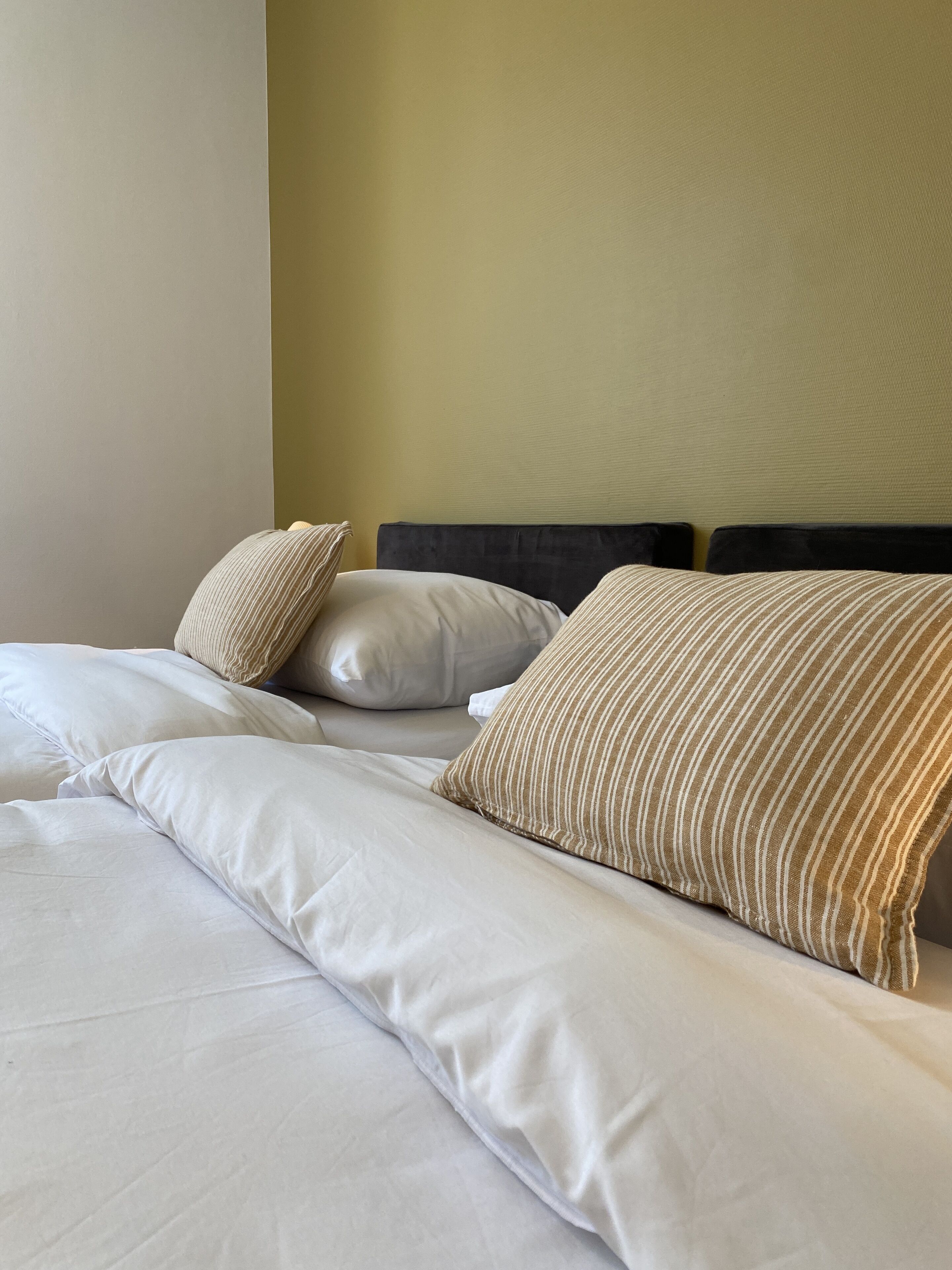 A neatly made bed with white linens and striped pillows, a dark headboard, and a plain wall.