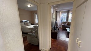 A bedroom with a bed, bedside table, and a TV. There is a living area with a sofa and a TV. The room has wooden flooring and white walls.