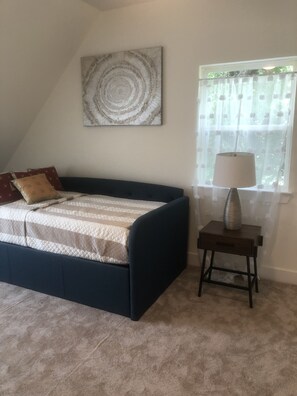 Une chambre avec un lit bleu foncé, une moquette beige, une œuvre d’art circulaire accrochée au mur, une fenêtre avec des rideaux légers et une table de chevet avec une lampe.