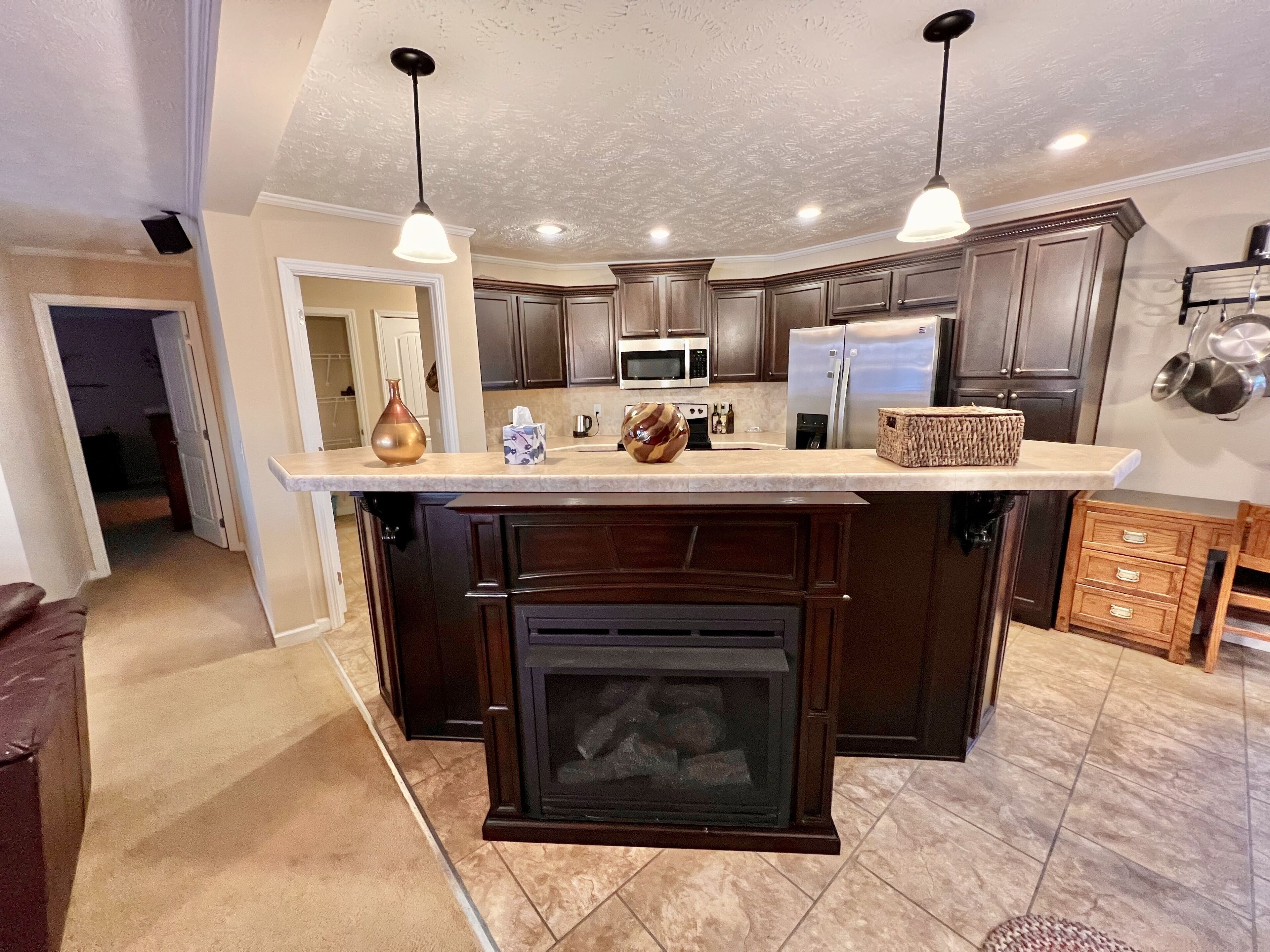 A modern kitchen with dark wood cabinets, a central island, a built-in fireplace, and pendant lighting.