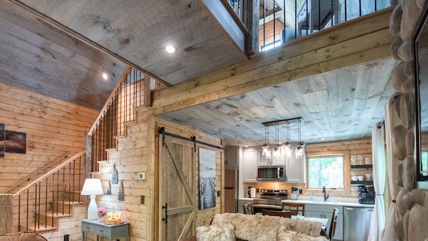 A rustic cabin interior with a wooden staircase, a cozy living area with a sofa and pillows, a kitchen with modern appliances, and a large window offering a view of the outdoors. - Treehouse, Hot Tub, Creekfront, Starlink Wi-Fi, Firepit (Franklin)
