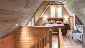 A cozy attic bedroom with a wooden sloped ceiling, a bed with multiple pillows, a nightstand, a chair, and a window providing natural light. - Treehouse, Hot Tub, Creekfront, Starlink Wi-Fi, Firepit (Franklin)