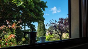A window view of a garden with a coffee maker and cup on the windowsill.
