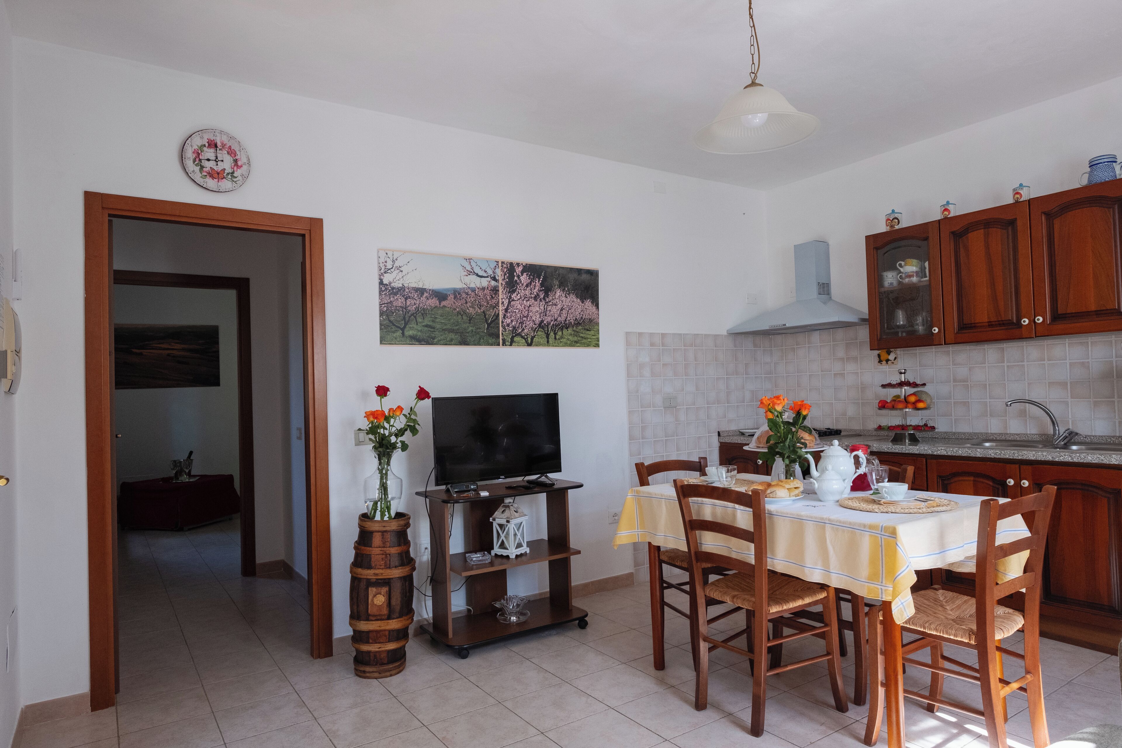 Una cocina con armarios de madera, una mesa de comedor con sillas, un televisor sobre un mueble bajo y un cuadro en la pared.