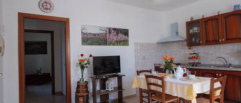 Una cocina con armarios de madera, una mesa de comedor con sillas, un televisor sobre un mueble bajo y un cuadro en la pared.
