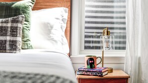 Cama bem arrumada com duas almofadas, mesa de cabeceira com caneca e livros, e janela com persianas.
