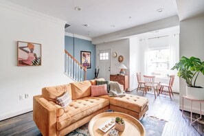 Una sala de estar moderna con un sofá modular, una zona de comedor con sillas de madera, una escalera y un amplio ventanal que proporciona luz natural.
