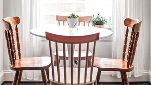 Uma área de refeições com uma mesa redonda e quatro cadeiras de madeira, uma janela com cortinas brancas e plantas em vasos no parapeito.