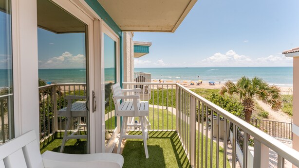 A balcony with white chairs and a table, overlooking a beach with people and umbrellas.