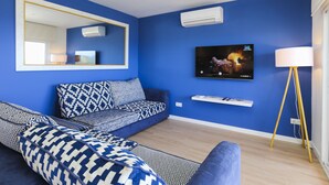 A modern living room with a blue wall, a sofa with patterned cushions, a television mounted above a shelf, and a floor lamp. - TOUS 2 Apartment - Only Families (Salou)