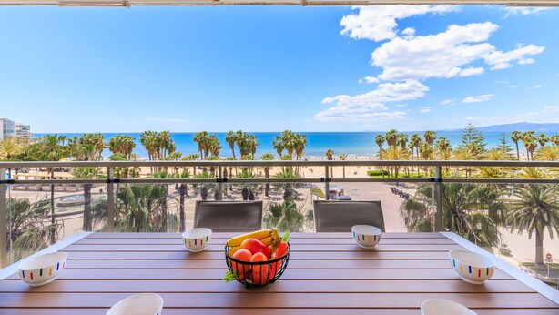A balcony with a wooden table, two chairs, and a bowl of fruit. The view includes a beach, palm trees, and the ocean. - TOUS 2 Apartment - Only Families (Salou)