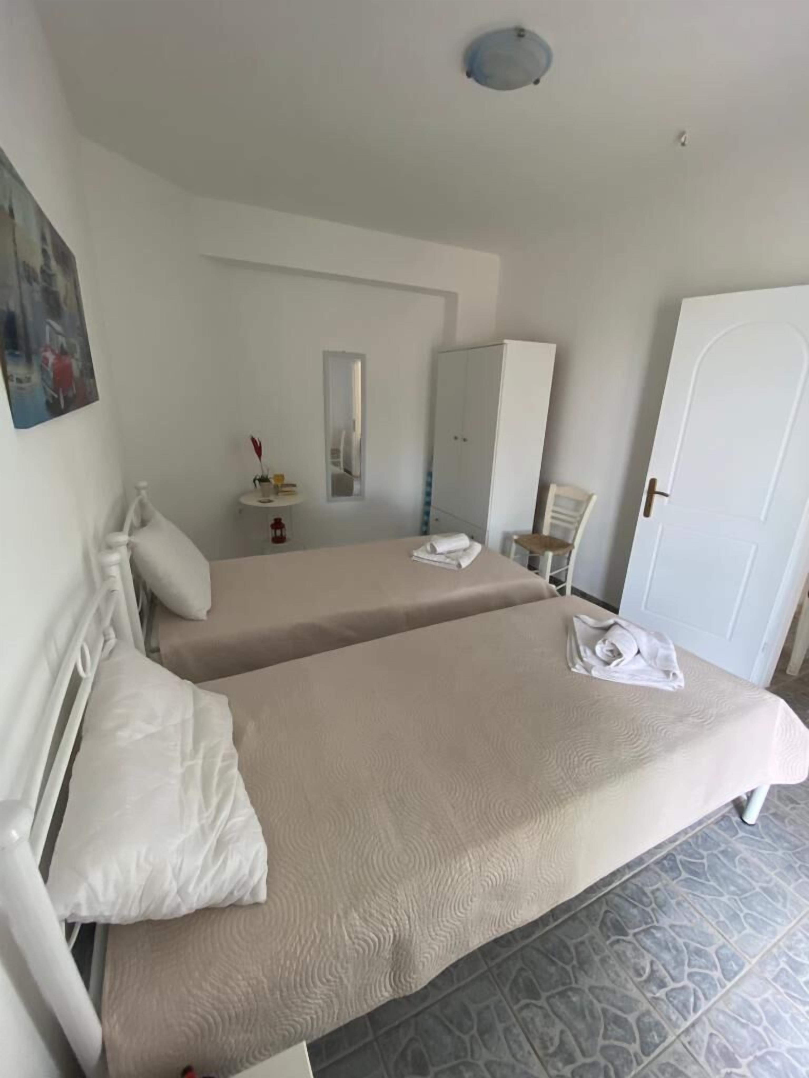 A hotel room with two beds, a wardrobe, a mirror, a small table with a lamp, and a door.