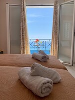 Habitación de hotel con una cama, dos toallas enrolladas, una mesita con un florero y vista al mar a través de una puerta corrediza de vidrio.