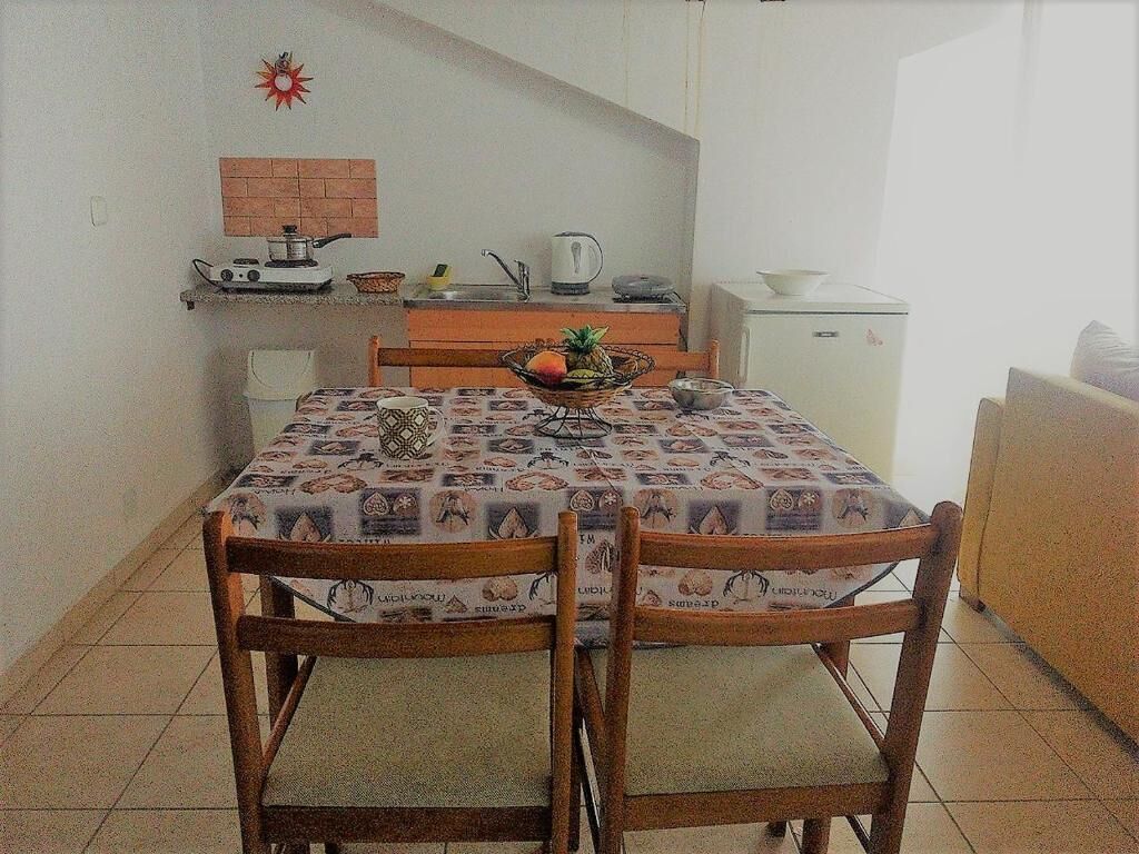 A dining area with a table covered in a patterned tablecloth, two wooden chairs, a kitchen area with a stove, kettle, and a refrigerator.