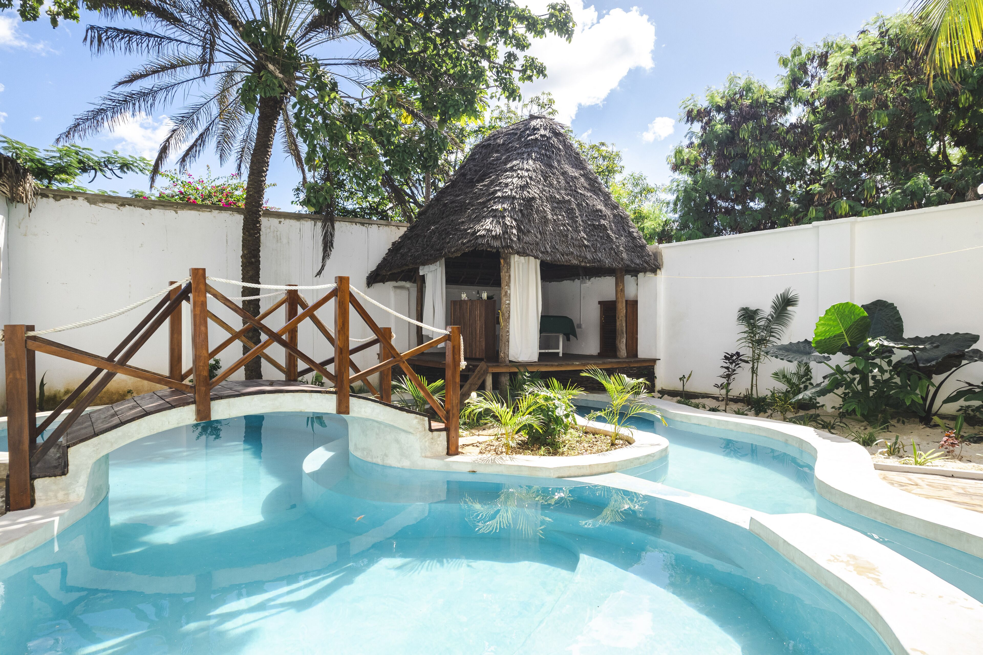 A swimming pool with a wooden bridge, a thatched-roof structure, and tropical plants.