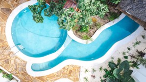 Una piscina con un design ondulato originale, circondata da piante diverse e una struttura a tetto di paglia.