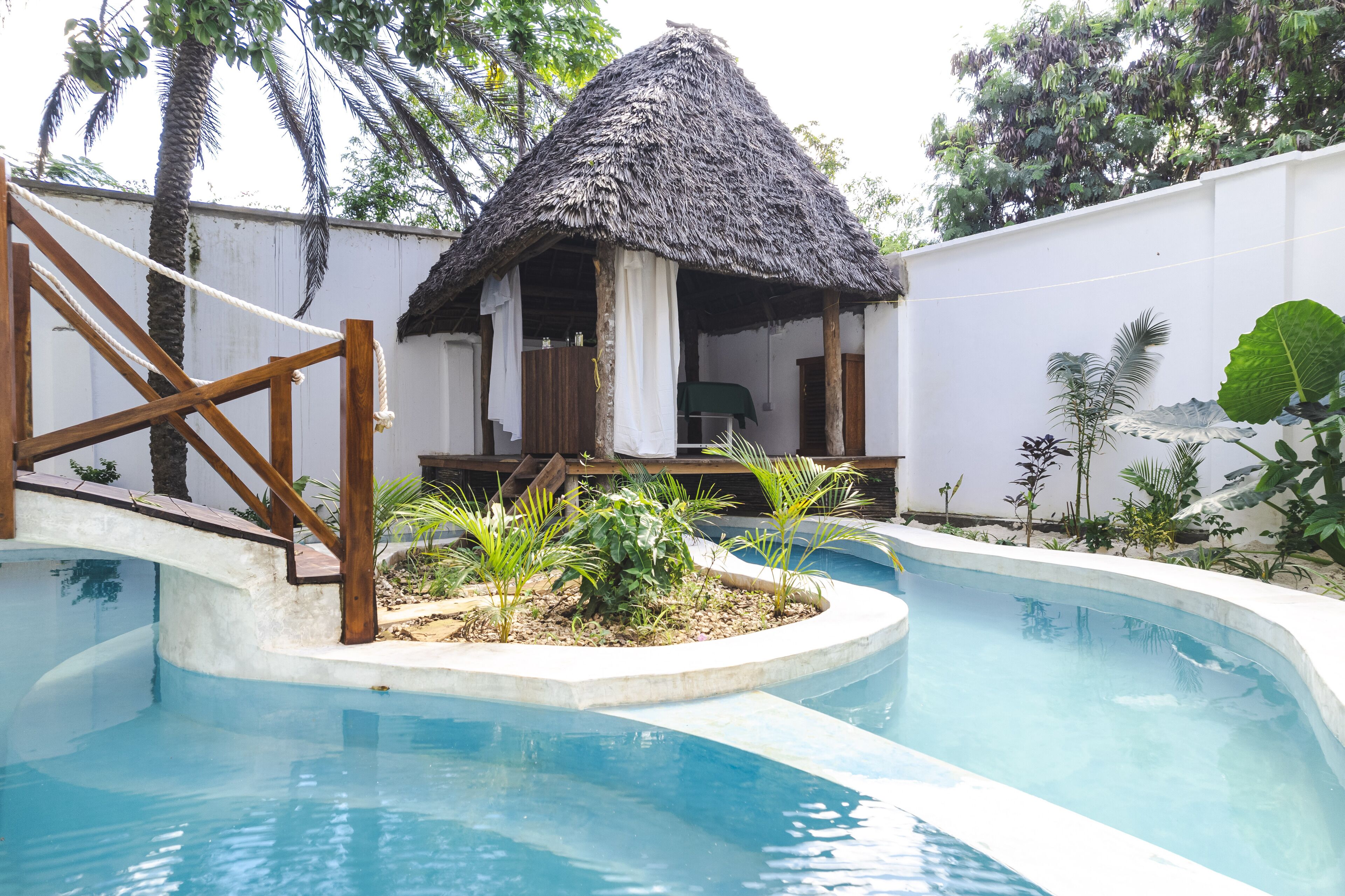 A pool area with a thatched-roof structure, wooden deck, and surrounding tropical plants.