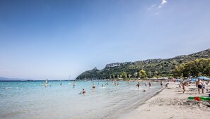 A beach with clear turquoise water, people swimming and sunbathing, and a rocky hillside in the background.