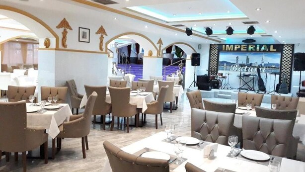 A restaurant with multiple dining tables and chairs, a stage with musical equipment, and a large screen displaying a cityscape. - Hotel Genatsvale Batumi (Batumi)