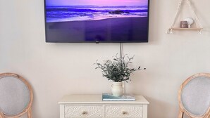Olohuoneessa on seinälle asennettu televisio, kaksi rottinkituolia, valkoinen konsolipöytä, jossa on kasvi, ja seinälle ripustettu makrame-seinävaate.