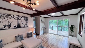 A spacious living room with a large sofa, a coffee table, and a rug. There is a painting on the wall, a lamp, and a large window with a view of trees.