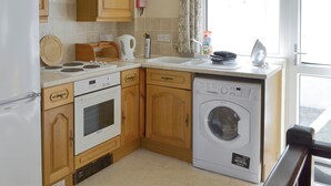 A compact kitchen with wooden cabinets, a built-in oven, a dishwasher, and a window with curtains. - Pirates Cove - UK44493 (Hope Cove, near Kingsbridge)